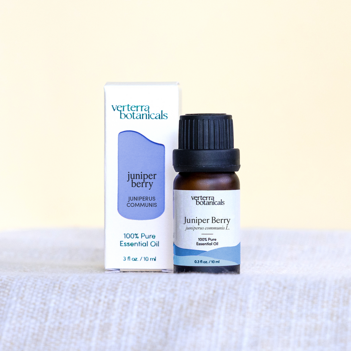 A bottle of Verterra Botanicals Juniper Berry essential oil next to its white and blue packaging box, displayed on a textured light gray fabric surface against a pale cream background.