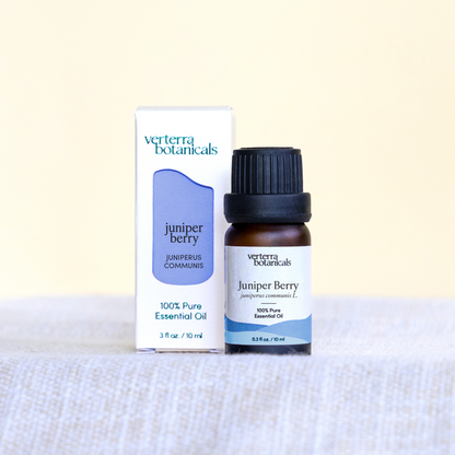 A bottle of Verterra Botanicals Juniper Berry essential oil next to its white and blue packaging box, displayed on a textured light gray fabric surface against a pale cream background.